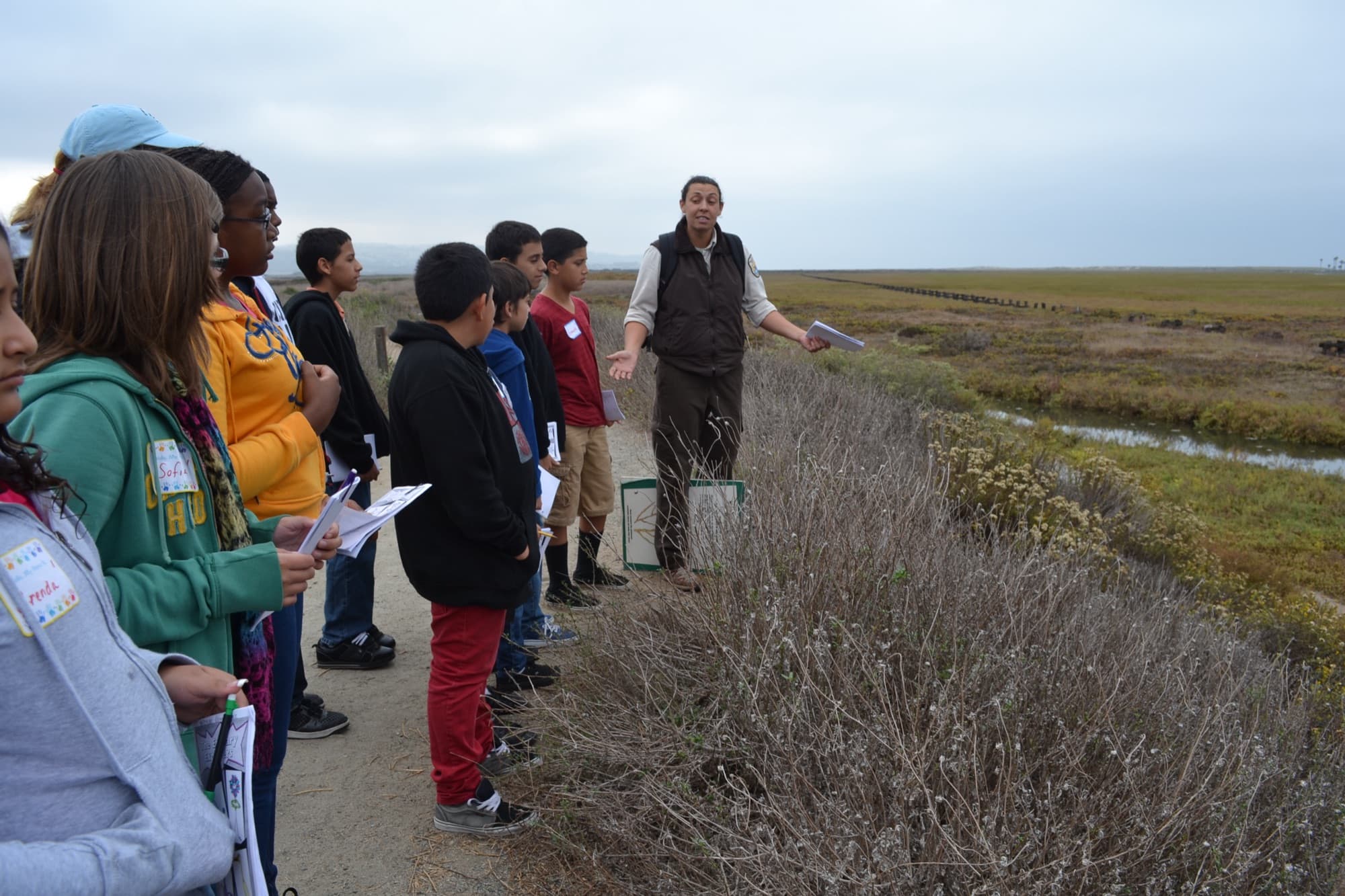 A group of middle school students holding worksheets listen to a wildlife refuge educator beside the salt marsh at the Tijuana Estuary