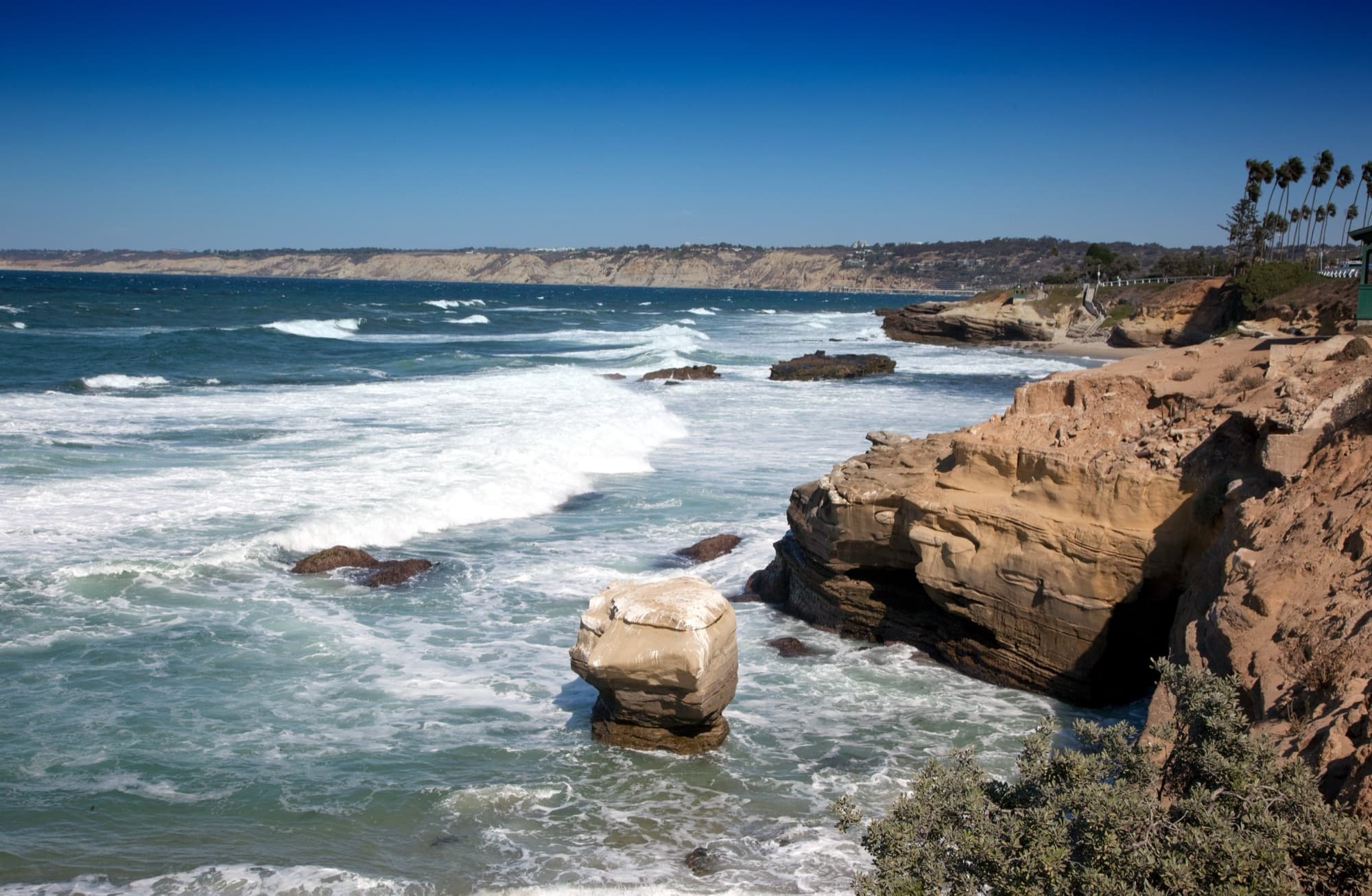 Bright blue waves breaking against sandstone cliffs and sea stacks along the La Jolla shoreline in San Diego