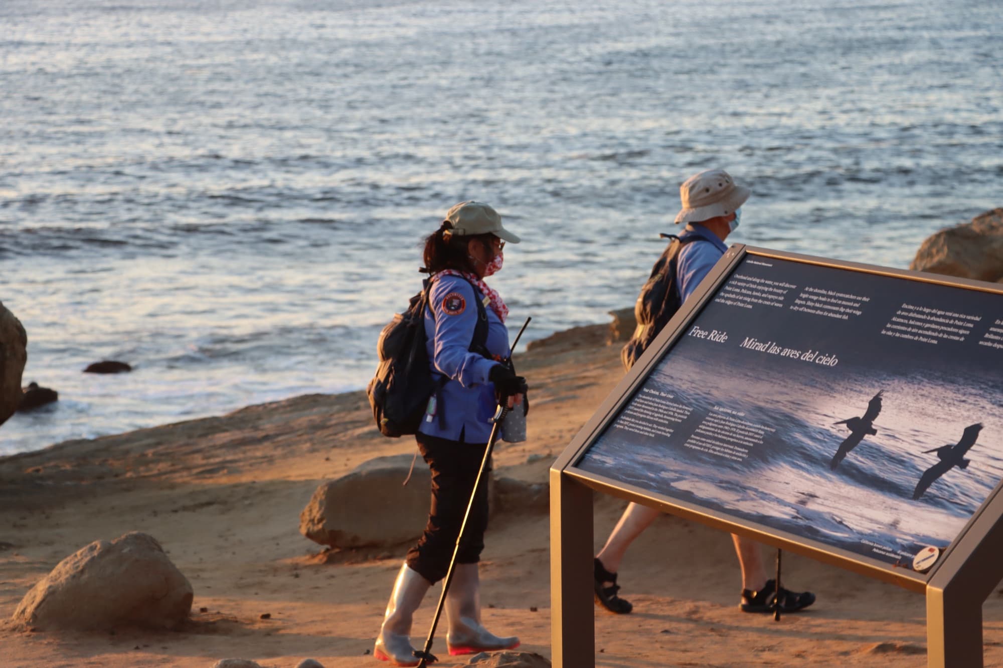 Two park volunteers walk past a shorebird interpretive sign beside the ocean at sunset at Cabrillo National Monument