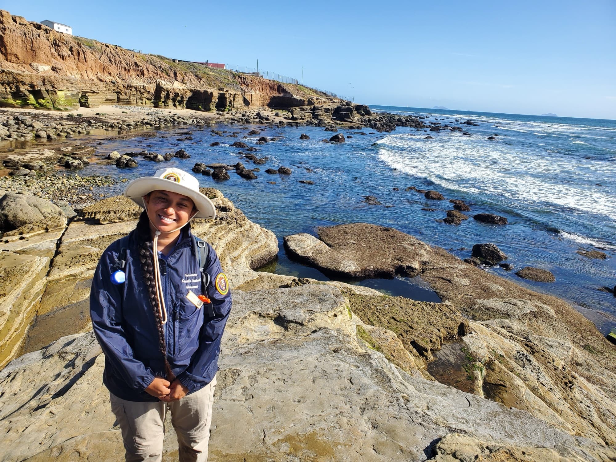 A smiling park volunteer in a uniform jacket and hat stands at the tide pool entrance at Cabrillo National Monument with waves behind